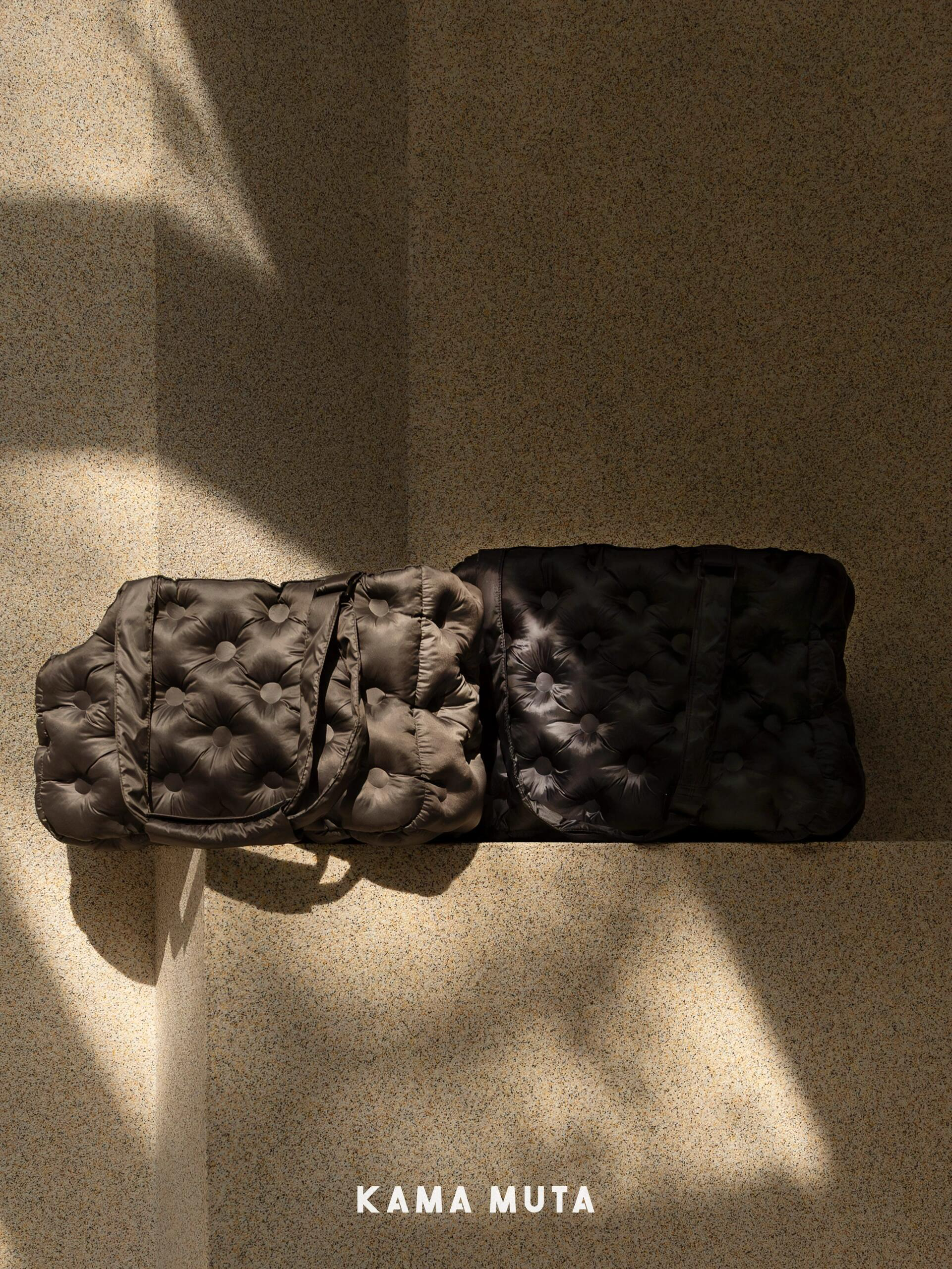 Dark quilted clutch bag on a textured beige surface with 'Kama Muta' branding.