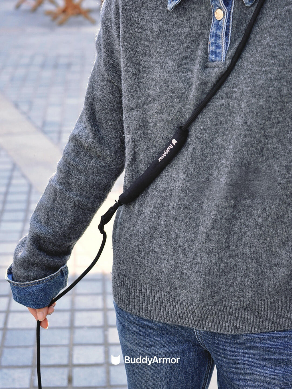 Person wearing a gray sweater with a black leash labeled 'Buddy Armor' on a blurred outdoor background.