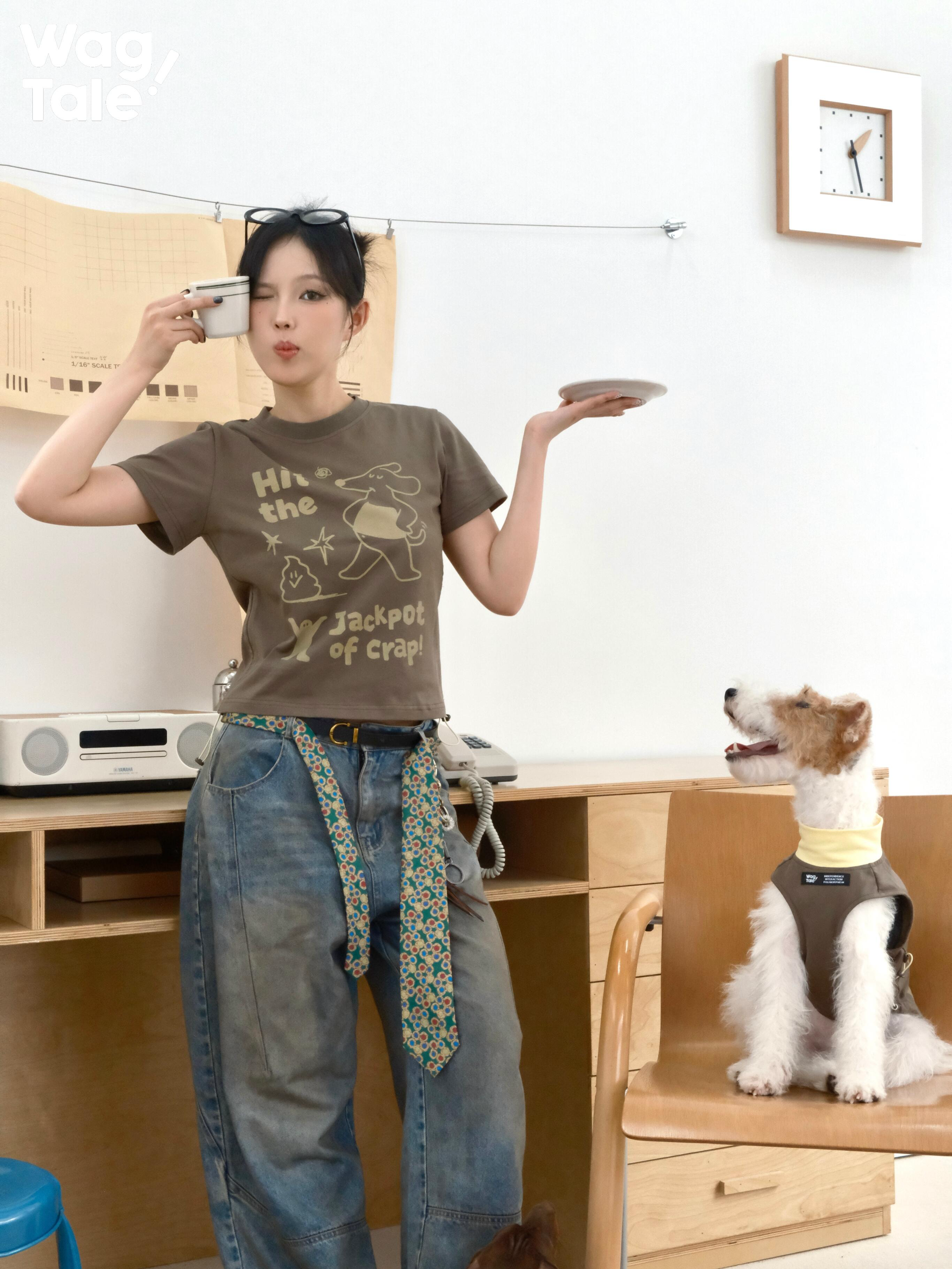 A person wearing a brown graphic cotton T-shirt poses with a cup and plate while a dog in a matching outfit sits on a chair, highlighting playful print styling.