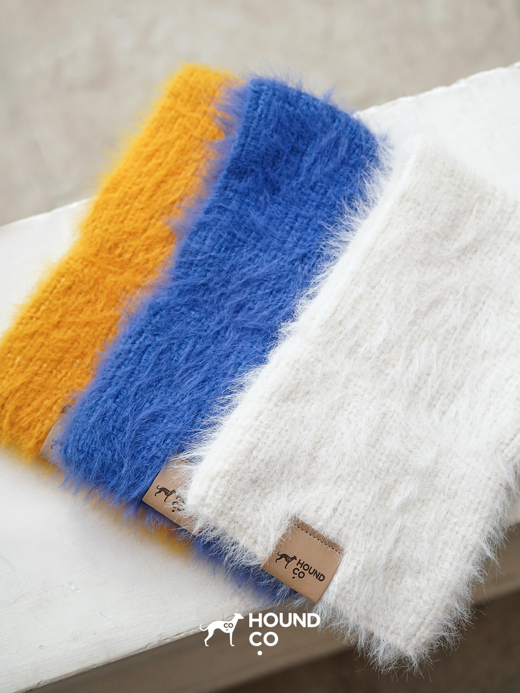 Hound-Co's collection of fuzzy knit snoods in mustard yellow, cobalt blue, and ivory, displayed together to showcase color options for sighthound owners.