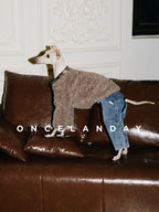 Italian Greyhound standing on a leather sofa wearing ONCELANDAY grey-brown lamb fleece dog jacket, showing textured fleece fabric in an indoor living room setting