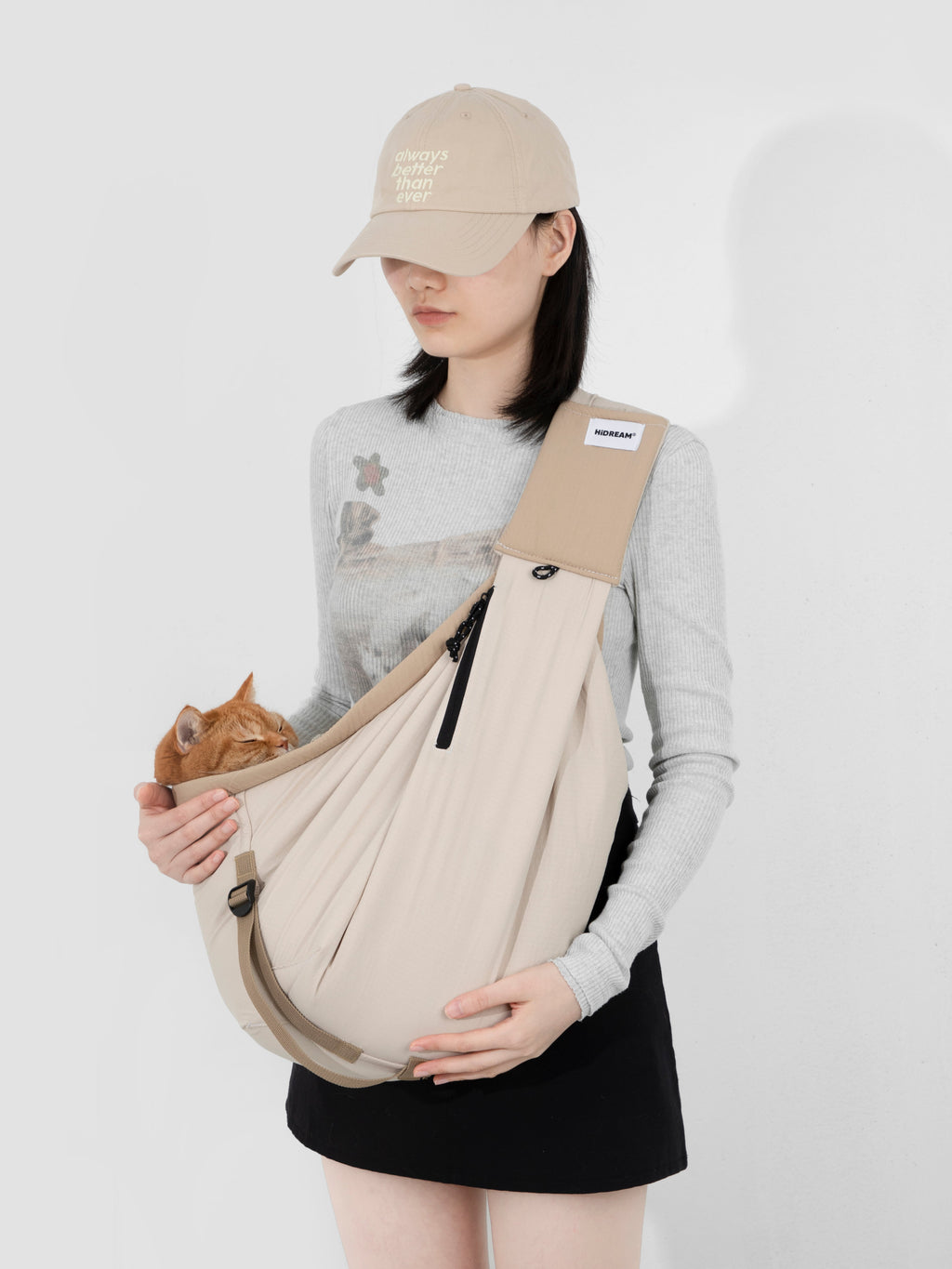 Woman wearing a beige pet sling carrier across the chest, holding an orange cat inside the soft fabric pouch, showing the diagonal carry position and close contact between pet and owner.