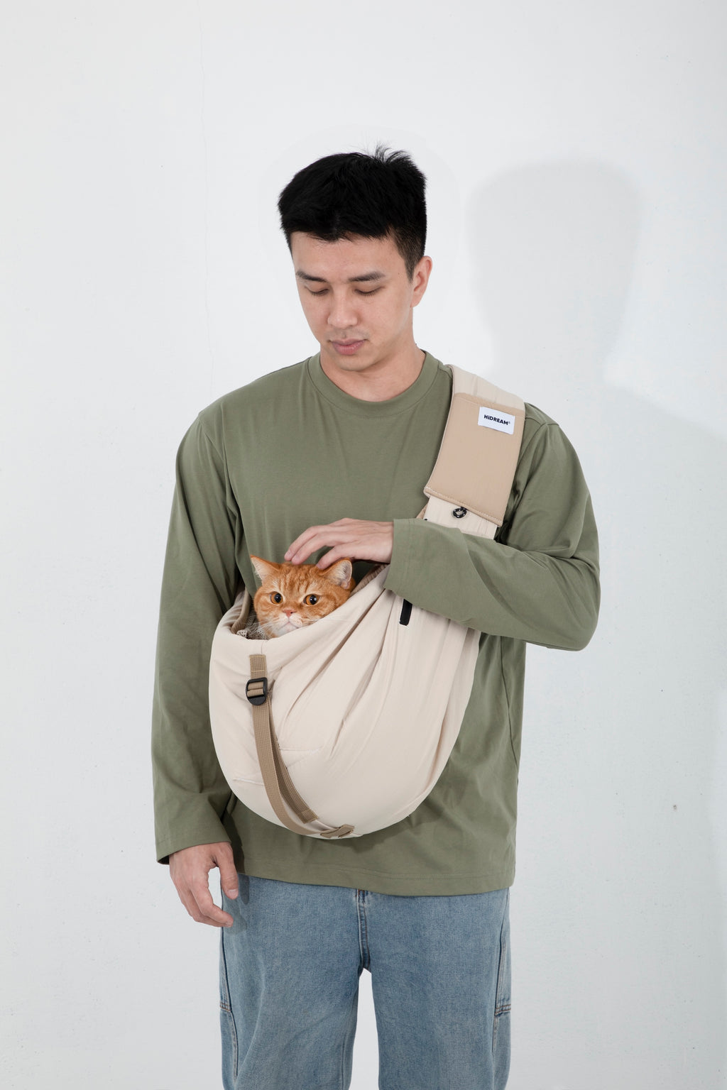 Man wearing a beige pet sling carrier on the front, gently holding an orange cat with its head visible above the opening, showing stable support and relaxed carry posture.