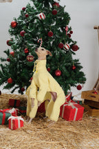 Dog wearing a yellow hooded dog jumpsuit standing upright beneath a decorated Christmas tree.